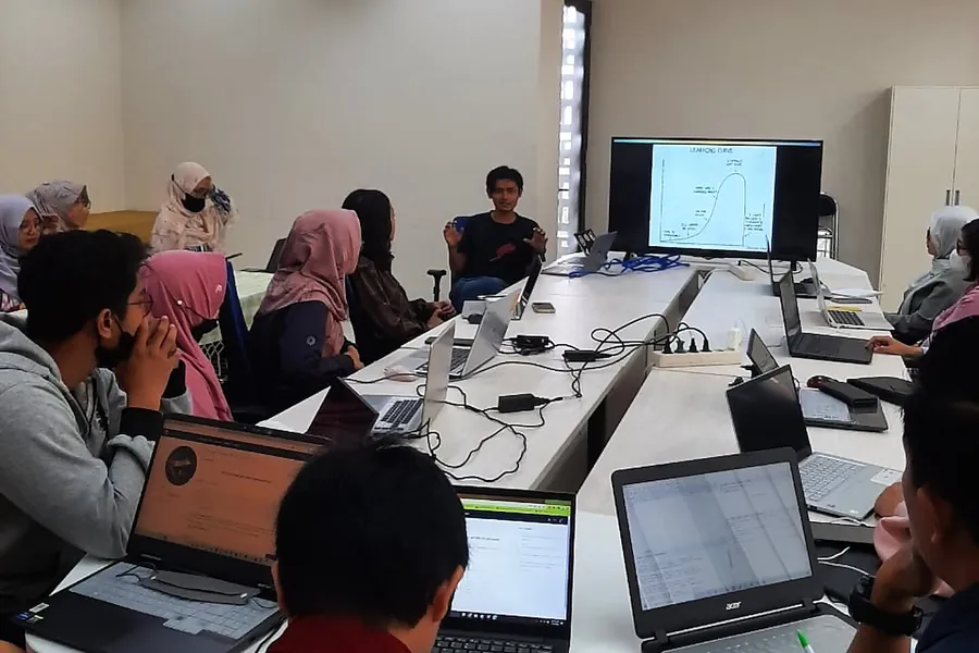 Students with laptops around a table during a coding workshop, with a learning curve slide on the screen